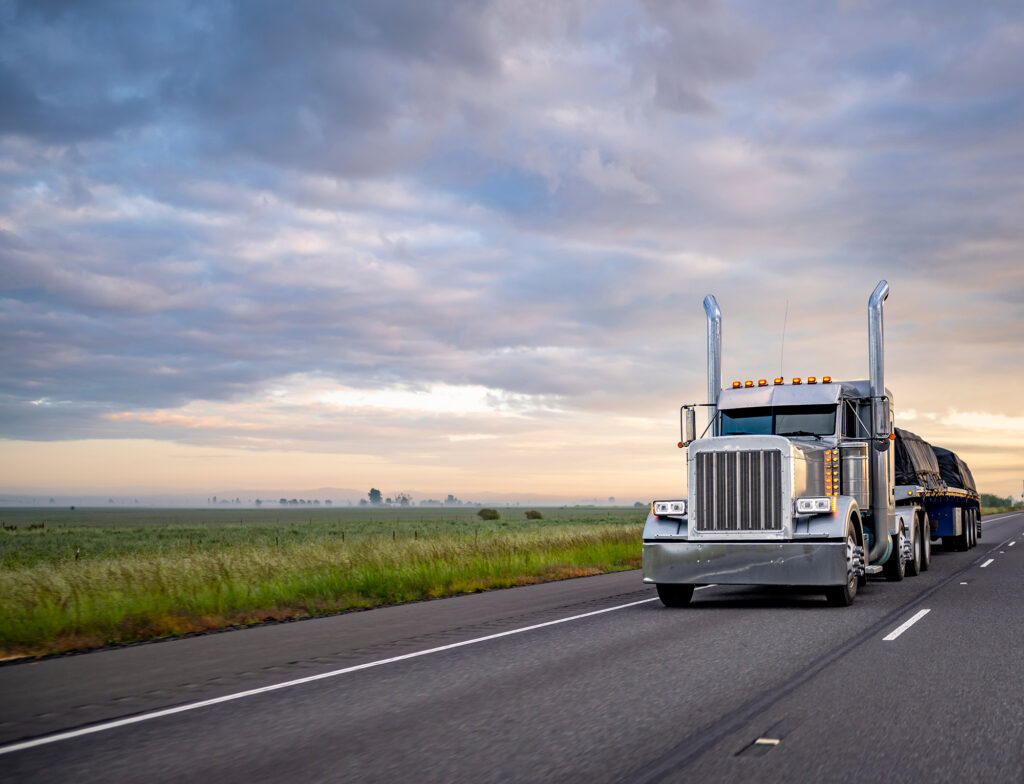 Gray classic big rig American semi truck with turned on lights and chrome exhaust pipes transporting covered cargo on flat bed semi trailer driving on the twilight highway road with stormy clouds sky